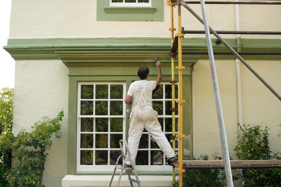 a man standing on a windowsill of a home reaching up with a paintbrush in his hand